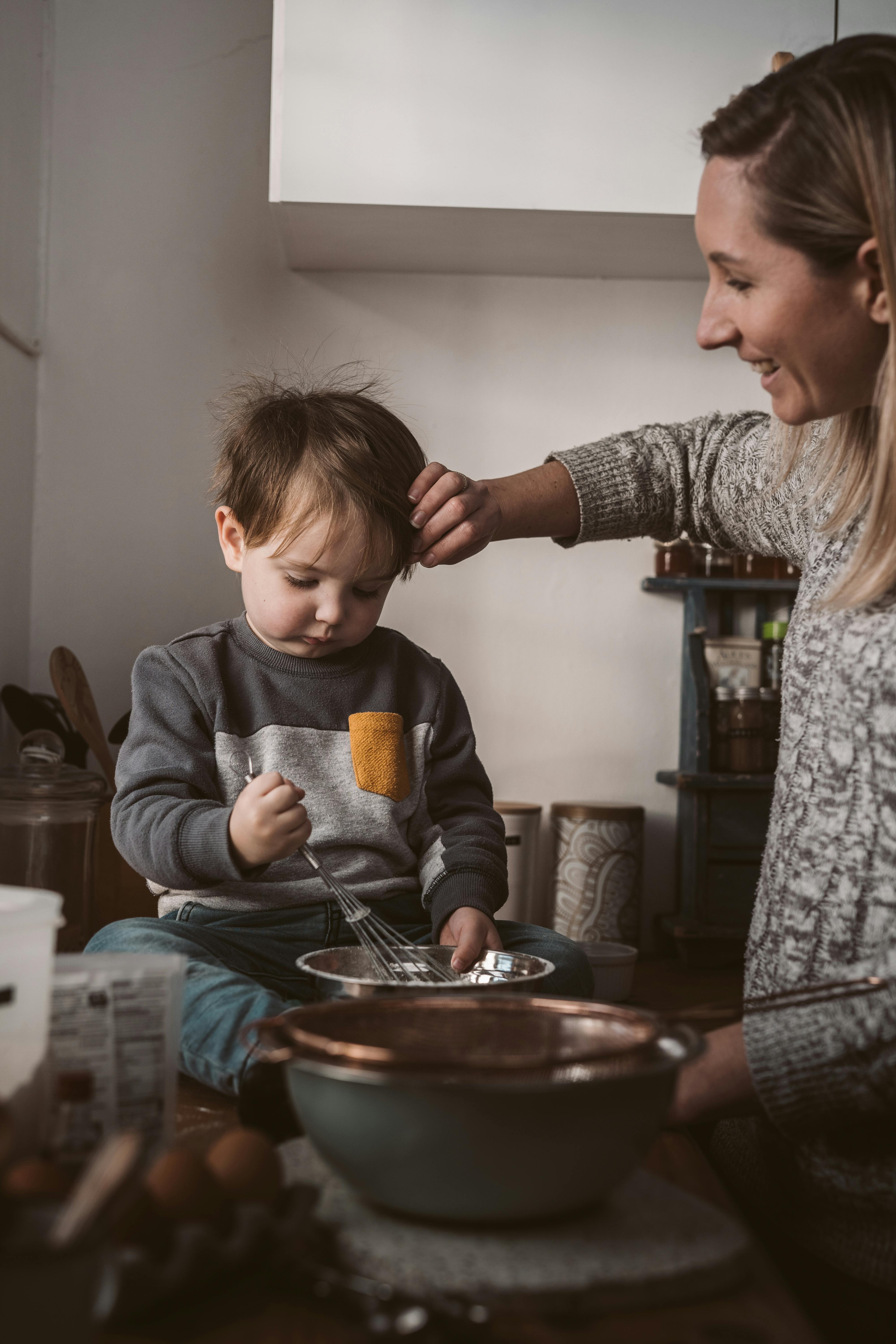 Photo of a mother and toddler. Toddler is sitting on the counter with mixing bowls with his mother beside him.
